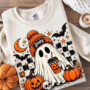Op de afbeelding: Een crèmekleurige sweater met ronde hals en een afbeelding van een witte geest die een oranje gebreide muts draagt met het woord Spooky erop. De geest houdt een oranje beker in de vorm van een pompoen vast, omringd door vleermuizen en sterren.