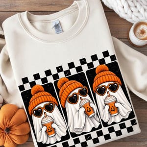 Peut inclure: Un sweat-shirt à col rond couleur crème avec une impression graphique de trois fantômes blancs portant des bonnets en tricot orange et des lunettes de soleil en forme de cœur. Chaque fantôme tient une boisson orange sur fond à damier.
