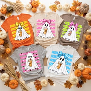 May include: Five t-shirts in orange, beige, brown, gray, and white, each featuring a graphic of a white ghost holding a coffee cup. Each ghost wears a different colored polka-dot bow, and the background of each graphic has horizontal stripes.