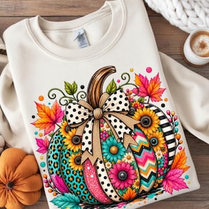 May include: A cream-colored crewneck sweatshirt featuring a large, colorful graphic of a pumpkin decorated with various patterns including leopard print, polka dots, stripes, and zigzags, accented with sunflowers and vibrant autumn leaves.