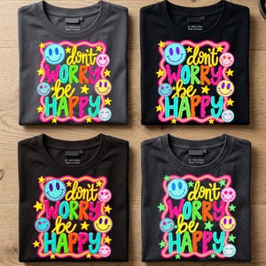 May include: Four t-shirts in black and dark gray featuring a colorful graphic print with the text "don't worry be HAPPY" surrounded by various smiley faces, stars, and a wavy border in bright shades of pink, yellow, blue, and orange.