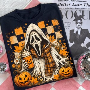 May include: A black t-shirt featuring a graphic of a ghost wearing a hooded cloak and a plaid scarf, holding a pumpkin-themed coffee cup. The design includes orange and black checkered patterns, autumn leaves, daisies, and two jack-o'-lanterns.