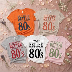 May include: Five short-sleeved t-shirts in orange, beige, gray, white, and light pink. Each shirt features the text "IT WAS BETTER IN THE 80's" printed in a bold, centered font. The shirts are arranged in a group on a light-colored fabric surface.