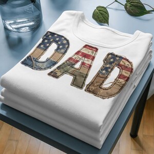 Könnte beinhalten: Ein weißes T-Shirt mit dem Wort DAD in großen, abgenutzten Buchstaben. Die Buchstaben haben ein strukturiertes Muster, das an die amerikanische Flagge mit blauen, weißen und roten Streifen und Sternen erinnert, sowie Camouflage- und Denim-Stoffmuster.