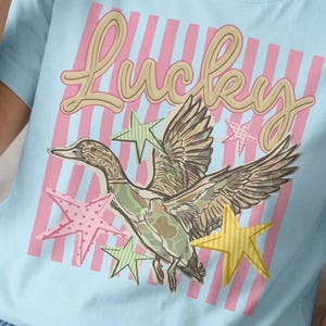 Puede incluir: Camiseta azul claro con un diseño de un pato con las alas extendidas, rodeado de estrellas. La palabra "Lucky" está escrita en cursiva sobre el pato. Rayas rosas y blancas en el fondo.