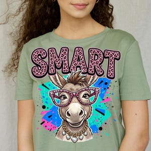 Op de afbeelding: Saliegroen T-shirt met een cartoon ezel met een roze bril en een gouden ketting. Het woord "SMART" staat bovenaan gedrukt in roze letters met luipaardprint. De ezel staat tegen een kleurrijke spetterachtergrond.