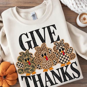 Op de afbeelding: Een crèmekleurig sweatshirt met ronde hals, zwarte tekst GIVE THANKS en drie cartoonkalkoenen. De kalkoenen hebben veren met camouflage-, luipaard- en zwart-wit geblokte patronen, elk met een bijpassende strik.