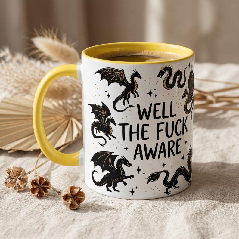 May include: White ceramic mug with a yellow handle and interior, filled with coffee. The mug features black dragon illustrations and the text "WELL THE FUCK AWARE".