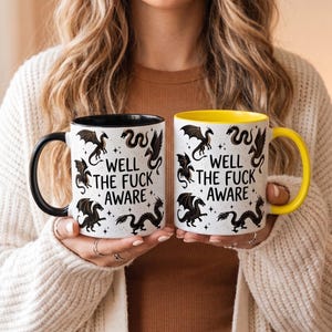 May include: Two white ceramic mugs with black and yellow accents. Each mug features a black dragon design and the text "WELL THE FUCK AWARE". The mugs are held by a person.