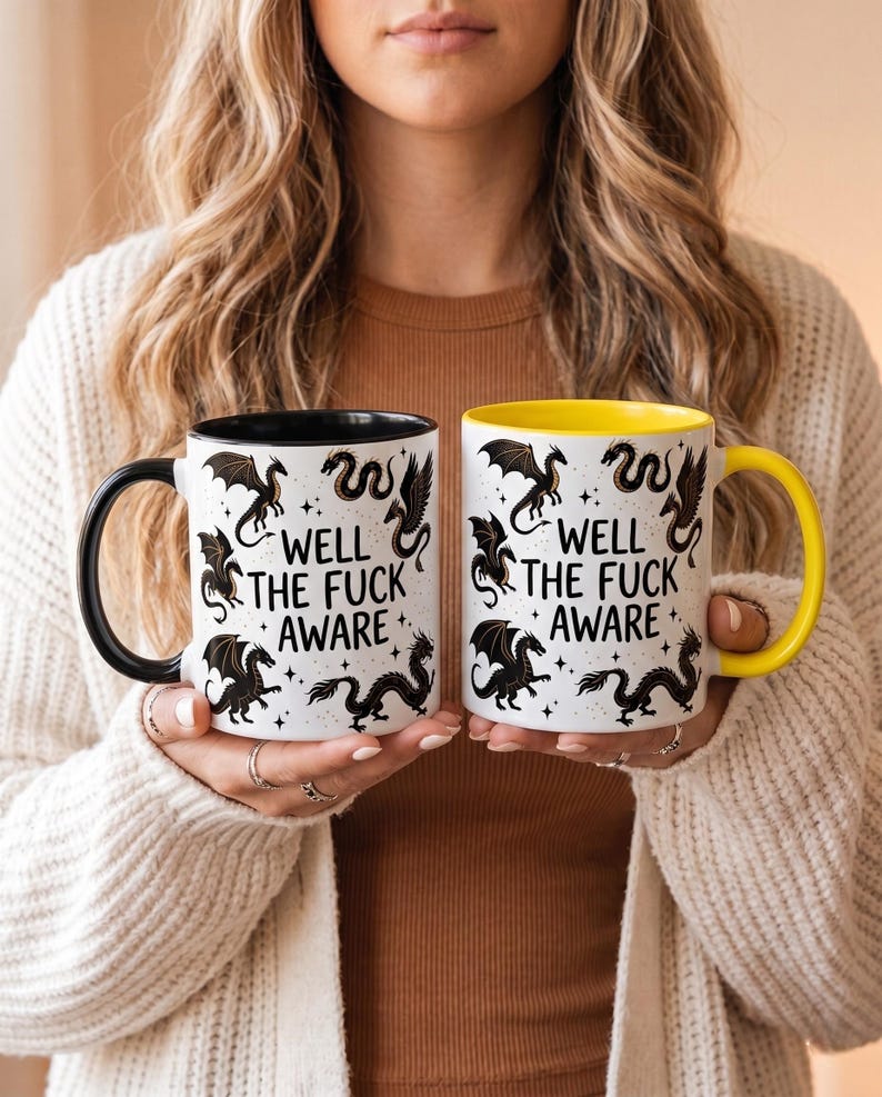 May include: Two white ceramic mugs with black and yellow accents. Each mug features a black dragon design and the text "WELL THE FUCK AWARE". The mugs are held by a person.