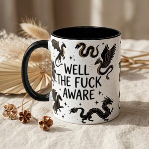 May include: White ceramic mug with a black interior and handle. The mug features black dragon illustrations and the text "WELL THE FUCK AWARE" in black. Gold stars and accents are scattered around the dragons.