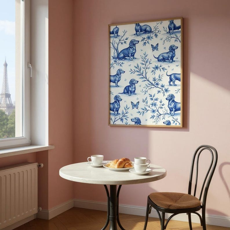 May include: A framed wall art print featuring a repeating pattern of blue dachshund dogs and butterflies on a white background with blue floral accents. The artwork is displayed on a pink wall above a small round table.