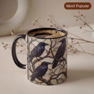 Può includere: Una tazza in ceramica con manico e bordo neri, caratterizzata da un motivo in stile vetrata con tre corvi neri appollaiati su rami marroni su sfondo crema. La tazza è riempita di caffè nero e presenta un'etichetta marrone Most Popular.