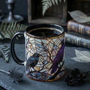 May include: A ceramic mug filled with coffee, featuring a stained-glass design of two crows perched on branches. The mug has a black handle and rim, with a gold trim. The background includes books and decorative elements.