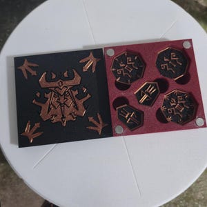 Op de afbeelding: Een set spelstukken met koperkleurige ontwerpen op zwarte en bordeauxrode achtergronden. De stukken zijn gerangschikt op een wit oppervlak, met extra losse stukken zichtbaar. De ontwerpen zijn gestileerde symbolen.