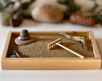 May include: A small Zen garden in a wooden tray. The garden features a bed of sand, a small wooden rake, a stack of stones, and a piece of driftwood. The tray is made of light-colored wood.