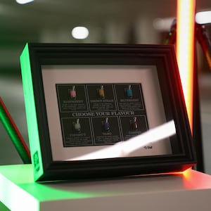 May include: A black framed display with a white background showing six small bottles with different coloured liquids. The bottles are labelled with the following flavours: Raspberry, White Peach, Blueberry, Coconut, Taro, and Coffee. The text "CHOOSE YOUR FLAVOUR" is printed below the bottles.