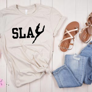May include: A light beige t-shirt with the word "SLAY" in bold black letters and a black silhouette of a dancer. The shirt is paired with blue denim jeans and brown sandals with gold straps. A bouquet of pink flowers is also present.