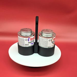 May include: Two bottles of Oatey PVC cement, one labeled "Purple Primer" and the other "Regular PVC Cement", are held in a black plastic holder with a handle. The holder is sitting on a white circular platform.