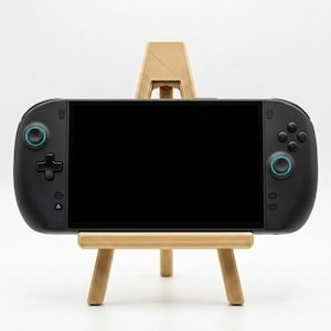 Pode incluir: Uma consola de jogos portátil preta com um grande ecrã retangular, um pad direcional e botões circulares com detalhes em azul-petróleo, apoiada num suporte de exposição bege de três pernas com um acabamento de superfície texturizado.