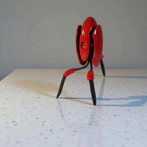 Può includere: Una statuetta di robot rosso e nero con una testa arrotondata e tre gambe. Il robot ha una finitura lucida e un singolo occhio arancione. Le gambe sono nere con dettagli rossi. La statuetta è su una superficie bianca maculata.