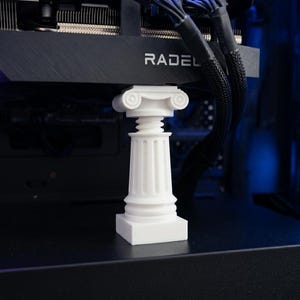 Könnte beinhalten: Eine kleine, weiße ionische Säule, die eine schwarze Computerkomponente trägt. Die Säule hat detaillierte Schnitzereien und eine quadratische Basis. Auf der Computerkomponente steht das Wort "RADEL". Der Hintergrund zeigt weitere Computerkomponenten und blaue Beleuchtung.