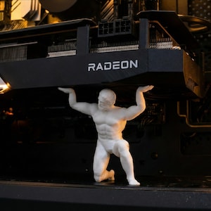 Puede incluir: Una figura blanca de un hombre musculoso sosteniendo una tarjeta gráfica Radeon negra dentro de una caja de computadora. La tarjeta gráfica tiene la palabra "RADEON" impresa. La figura está arrodillada, con los brazos levantados para sostener la tarjeta.