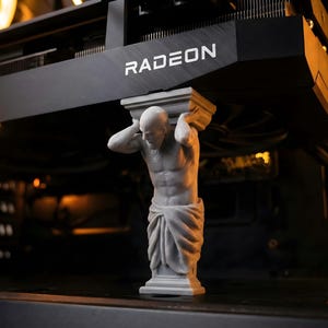 Könnte beinhalten: Eine graue Statue eines Mannes, der eine schwarze Radeon-Grafikkarte stützt. Die Statue ist ein dekoratives Computerzubehör, das einem Gaming-PC-Build eine einzigartige Ästhetik verleiht. Die Radeon-Karte trägt den Markennamen in Weiß.