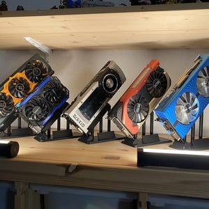 May include: A display of five colorful graphics cards, each mounted on a black stand. The cards feature various color schemes, including blue, orange, and red, with visible fans and heat sinks. The cards are arranged on a wooden shelf.