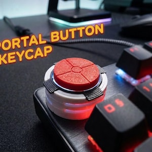 May include: A circular mechanical keyboard keycap featuring a red top with a cross-shaped indentation, set on a white base with black accents. The text PORTAL BUTTON KEYCAP is visible in orange letters above the keycap on a black keyboard.