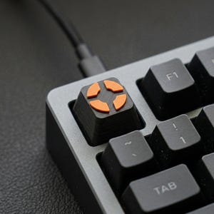 May include: Close-up of a mechanical keyboard with a custom keycap. The keycap is black with an orange design. Other black keys are visible, along with a gray keyboard frame and a black cable.
