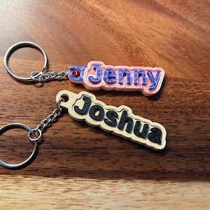 Puede incluir: Dos llaveros personalizados con nombres. Uno dice "Jenny" en letras azules con contorno rosa, y el otro dice "Joshua" en letras negras con contorno beige. Ambos llaveros tienen anillas plateadas.