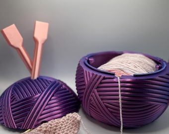May include: A purple yarn bowl with a pink ball of yarn inside. The yarn bowl has a decorative pattern and a pink yarn tail is coming out of the bowl. There is a pink crochet hook and a pink crocheted piece of fabric next to the bowl.