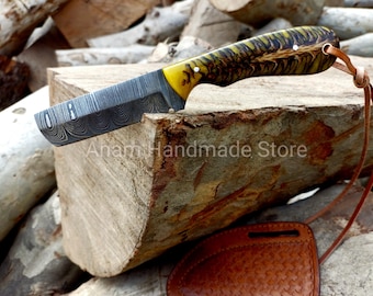 May include: A handmade knife with a patterned steel blade and a yellow and brown handle. The knife rests on a wooden block, with a leather sheath and strap visible. The text "Anam Handmade Store" is printed on the wood.