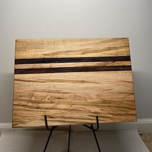 Könnte beinhalten: Ein großes Schneidebrett aus Holz mit einer natürlichen Holzoptik und zwei schwarzen Streifen. Das Brett steht auf einem schwarzen Metallständer.