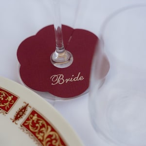 Może przedstawiać: Bordowa, kwiatowa wizyt&oacute;wka z napisem "Bride" w eleganckiej czcionce. Kieliszek do wina spoczywa na karcie, z dekoracyjnym talerzem na pierwszym planie. Talerz ma białe tło z ozdobną czerwoną i złotą obw&oacute;dką.