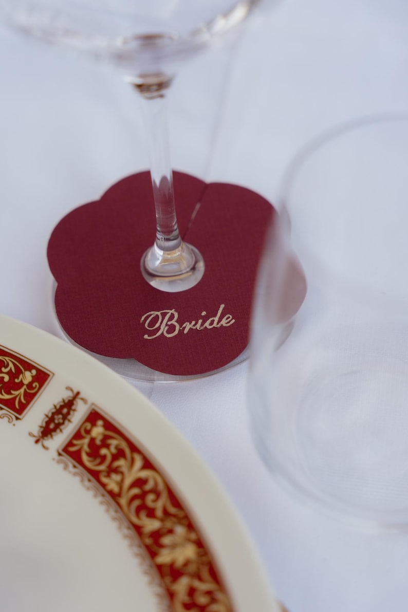 Może przedstawiać: Bordowa, kwiatowa wizyt&oacute;wka z napisem "Bride" w eleganckiej czcionce. Kieliszek do wina spoczywa na karcie, z dekoracyjnym talerzem na pierwszym planie. Talerz ma białe tło z ozdobną czerwoną i złotą obw&oacute;dką.