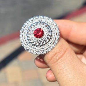 May include: A silver-toned ring with a large, round design. The ring features a central, vibrant red gemstone surrounded by concentric circles of intricate filigree work. The ring is held by a hand, showcasing its ornate details.