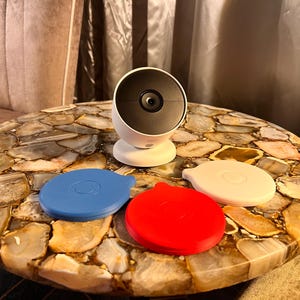May include: A white security camera with a black lens sits on a table with a brown and white agate surface. Three silicone covers in blue, red, and white are also on the table.