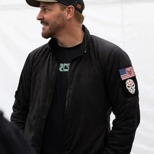 Könnte beinhalten: Schwarze Bomberjacke mit einem Aufnäher mit amerikanischer Flagge und Totenkopf-Design. Die Jacke wird mit einer grün-schwarzen Baseballkappe getragen. Die Person trägt ein schwarzes T-Shirt mit den grünen Buchstaben "RG".