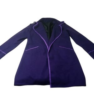 Puede incluir: Un abrigo largo de color púrpura con ribetes púrpuras en los bordes, bolsillos y puños. El abrigo tiene un diseño clásico con cuello de solapa y forro interior oscuro. El abrigo está sobre un fondo blanco.