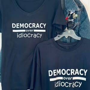 Peut inclure: Deux t-shirts bleu marine avec l'inscription blanche "DEMOCRACY over idiocracy". L'un a un col en V, l'autre un col rond. Une veste en jean est visible en arrière-plan.