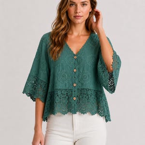 May include: A dark teal button-down blouse featuring a V-neckline, three-quarter length sleeves, and intricate circular crochet lace detailing across the chest and hemline. The garment is paired with plain white trousers.
