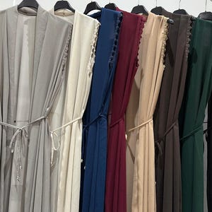Può includere: Una fila di abiti senza maniche in vari colori, tra cui grigio, crema, blu navy, bordeaux, beige, marrone, verde scuro e nero. Ogni abito ha una cintura abbinata e un bordo in pizzo sulle spalle. Gli abiti sono appesi a grucce nere.