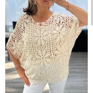 May include: Cream-coloured crochet top with a floral design. The top has a round neckline and short sleeves. The crochet pattern creates a delicate, openwork texture. The top is paired with white trousers.