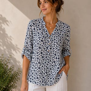 May include: A long-sleeved blouse featuring a white base with a dense pattern of small dark blue flowers. The shirt has a V-neckline with a partial button-down front, light-coloured buttons, and sleeves that are rolled up to the elbows.