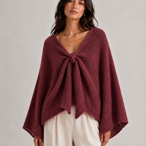 May include: A deep burgundy knit poncho featuring a V-neckline and a prominent front tie detail. The garment has wide, flared sleeves and a textured, fuzzy yarn construction that drapes loosely over the torso, paired with light cream trousers.