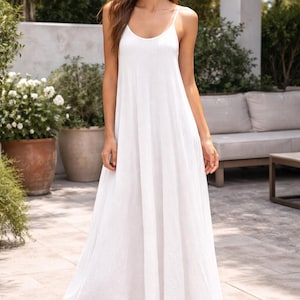 May include: A white maxi dress with spaghetti straps. The dress has a flowing, A-line silhouette. The model is wearing nude heels. The background includes greenery and a patio setting.