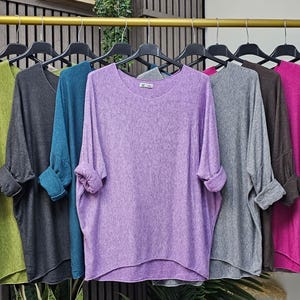 Könnte beinhalten: Eine Kollektion langärmliger Oberteile in verschiedenen Farben, darunter Marineblau, Beige, Grün, Anthrazit, Türkis, Lavendel, Grau, Braun, Fuchsia und Schwarz. Die Oberteile hängen auf einem goldenen Kleiderständer und präsentieren ihre verschiedenen Farbtöne und Stile.