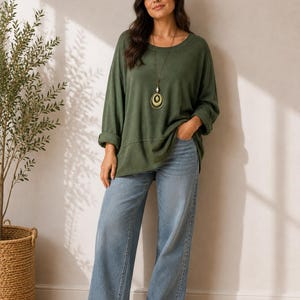 May include: An olive green long-sleeved top with a relaxed fit and a rounded neckline, paired with light blue wide-leg denim jeans and white sneakers. A long necklace with a large circular pendant hangs around the neck over the green top.